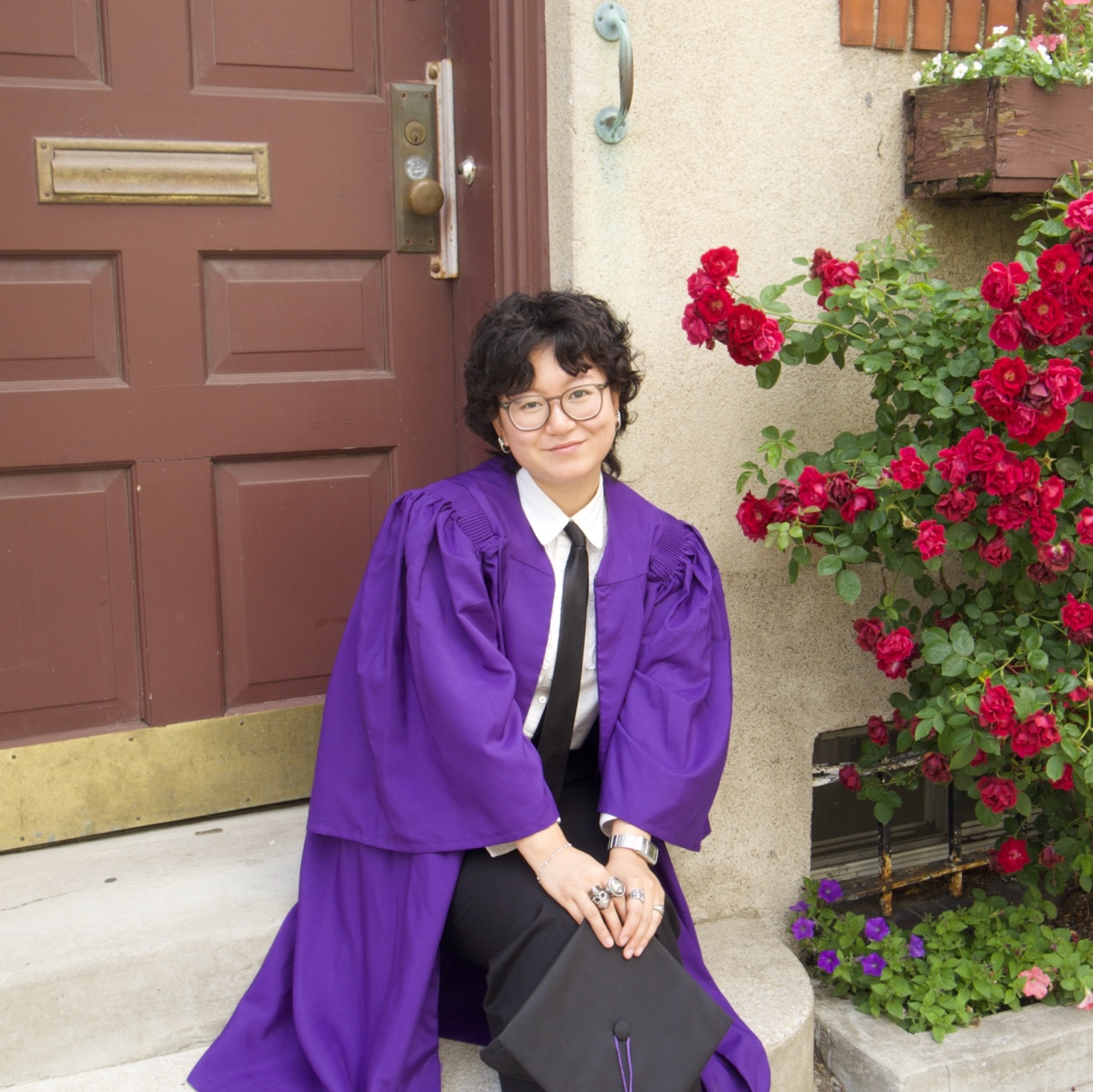 Charlene Chow smiling outside in her graduation gown, holding her cap, next to a rose bush